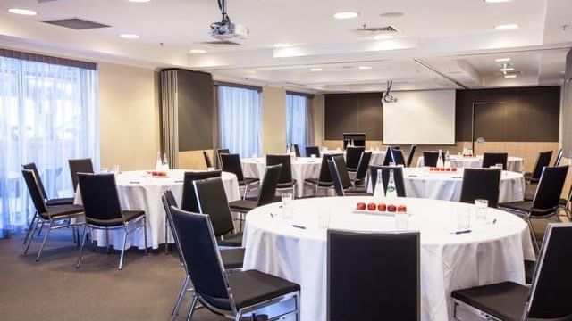 Bright conference room set with multiple round tables, black chairs, and a large projector screen at The Sebel Brisbane
