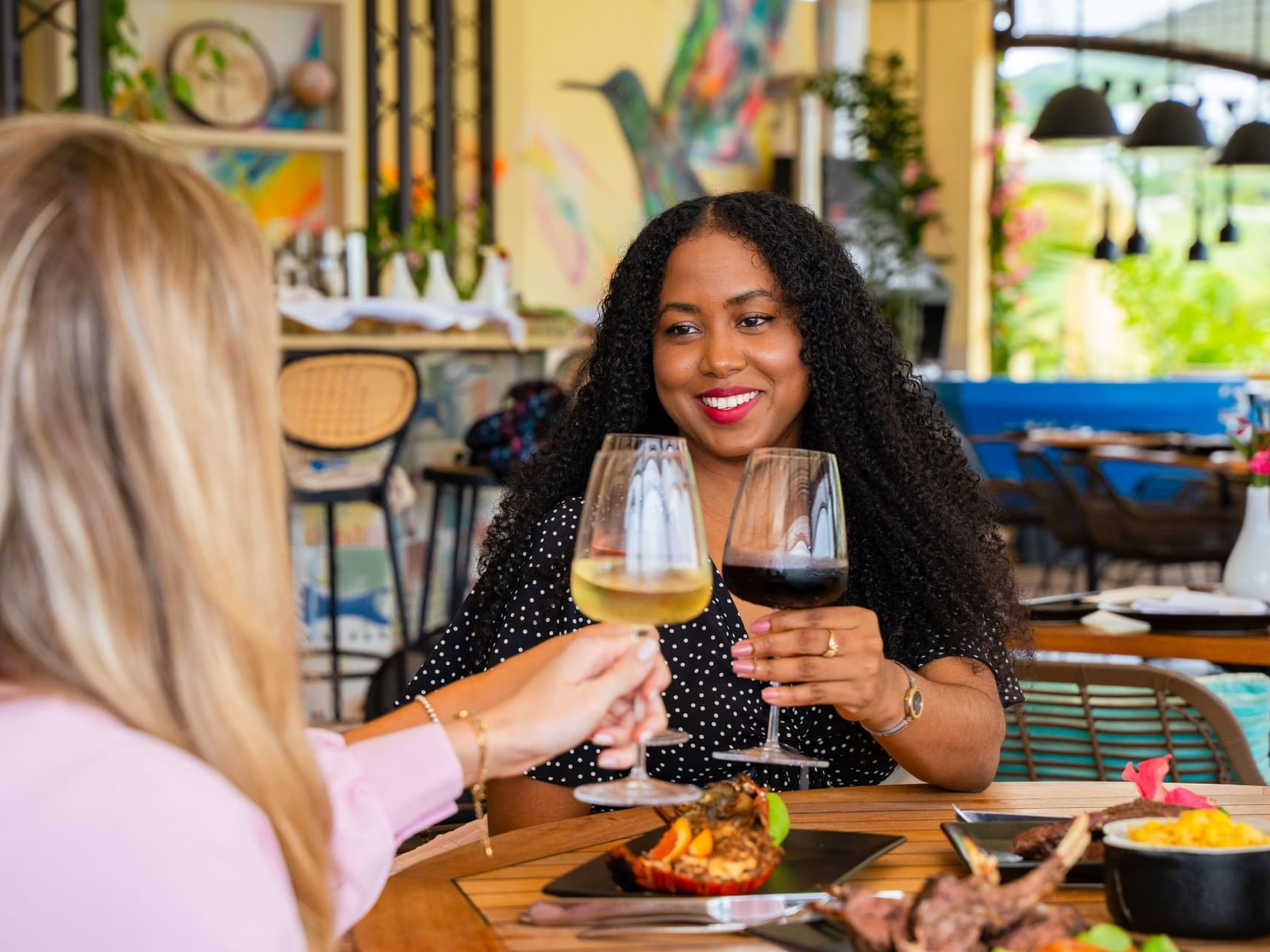 Twee vrouwen proosten met glazen wijn in Bobbie’s Beach Club, met gerechten op een houten tafel bij Golden Rock Resort