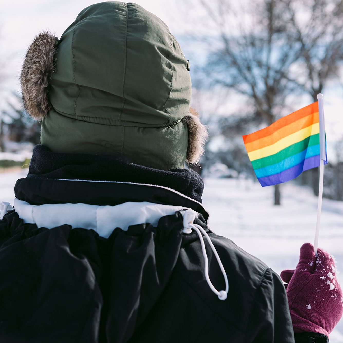 Person in winter coat holding a rainbow pride flag in a snowy outdoor setting for ADK Winter Pride.