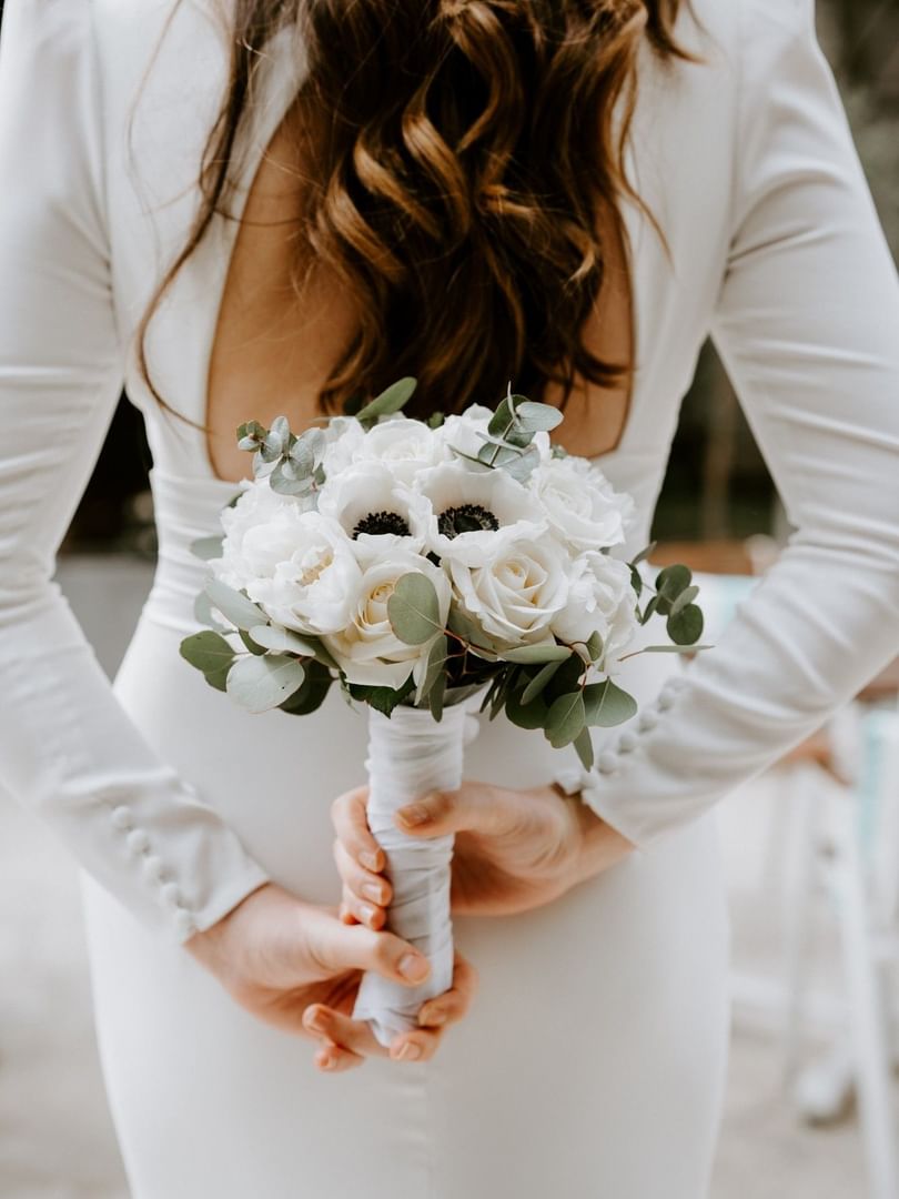 Bride in elegant white gown holding white rose and anemone bouquet with eucalyptus accents outdoors