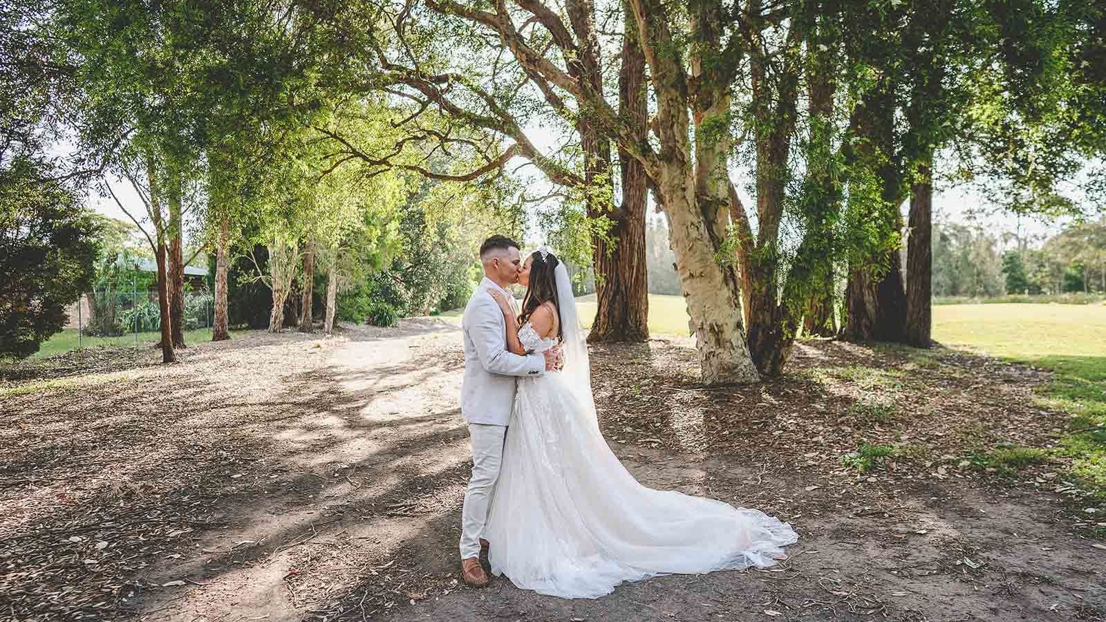 A couple in wedding attire embracing under a tree canopy at Mercure Kooindah Waters