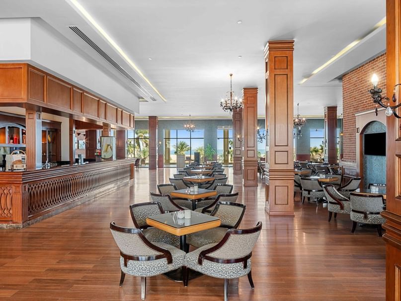 A spacious dining area with wooden floors, tables, chairs, and a counter, illuminated by chandeliers and wall sconces.