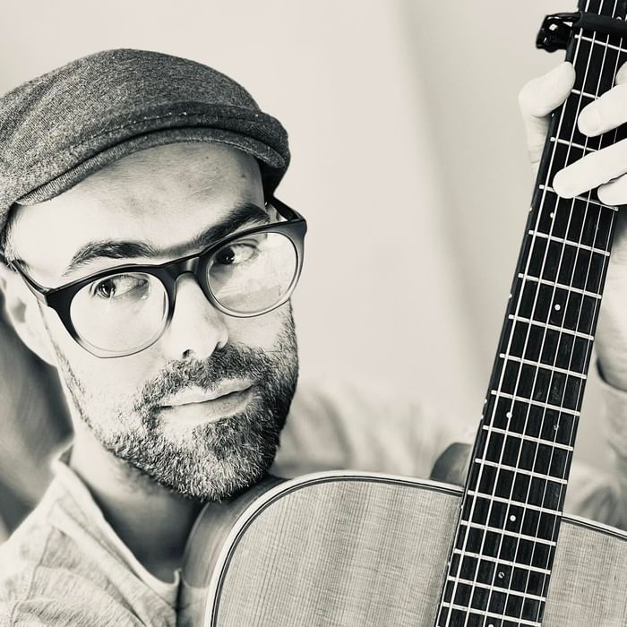 Black and white portrait of Michael Sigouin in cap and glasses holding acoustic guitar neck at Rodd Brudenell River Resort