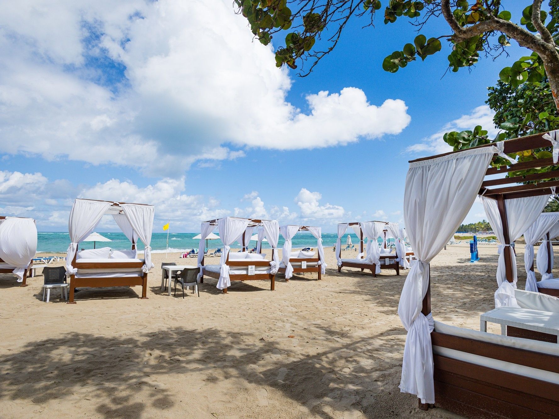 White-draped cabanas line a pristine sandy beach with turquoise waters under a bright blue sky with fluffy clouds.