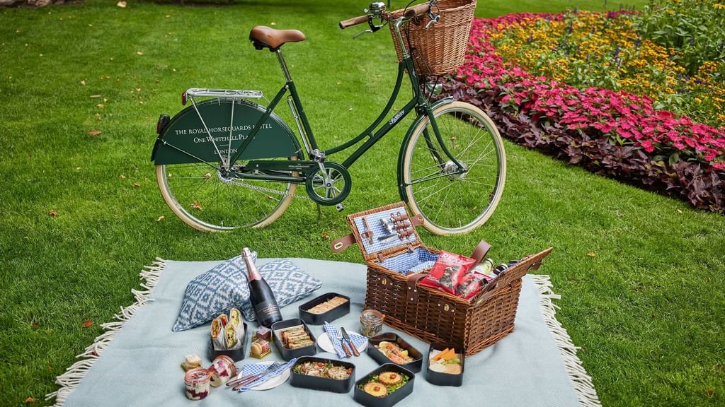 Picnic blanket with assorted food, wine, and a bicycle in a park with colorful flowers.