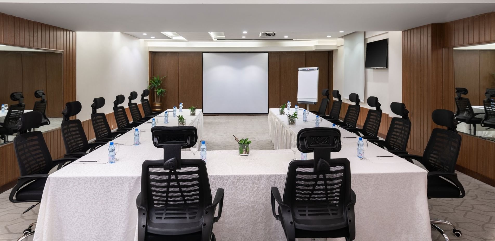 U-shaped table with water bottles by black mesh chairs facing the projector screen in Meeting Room A at Warwick Hotel Jeddah