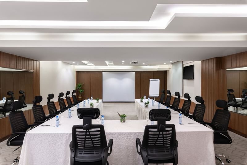 U-shaped table with water bottles by black mesh chairs facing the projector screen in Meeting Room A at Warwick Hotel Jeddah