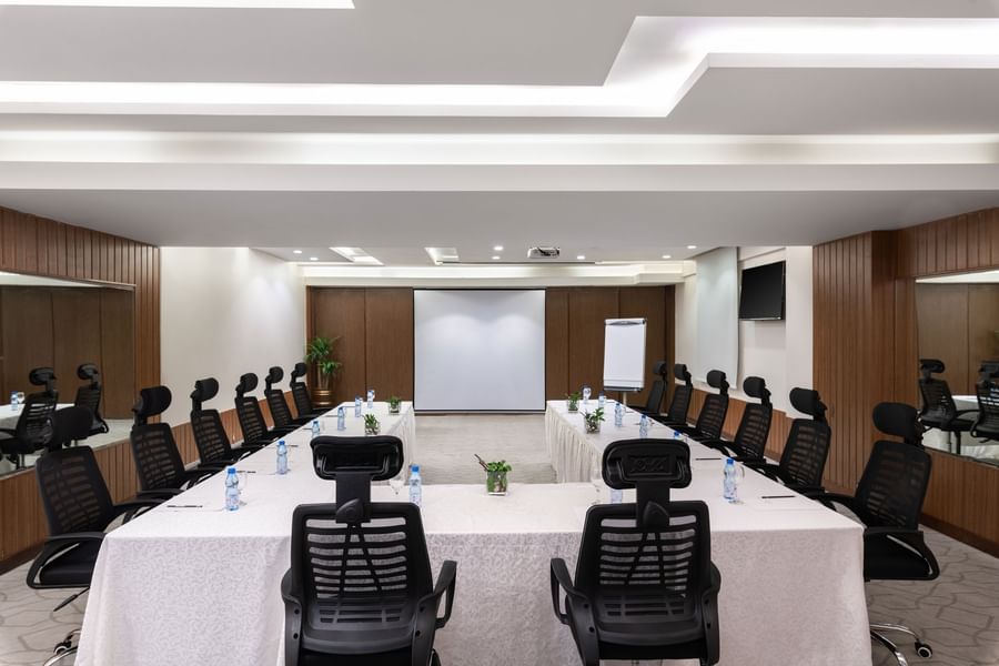 U-shaped table with black mesh chairs facing the projector screen in Meeting Room A at Warwick Hotel Jeddah