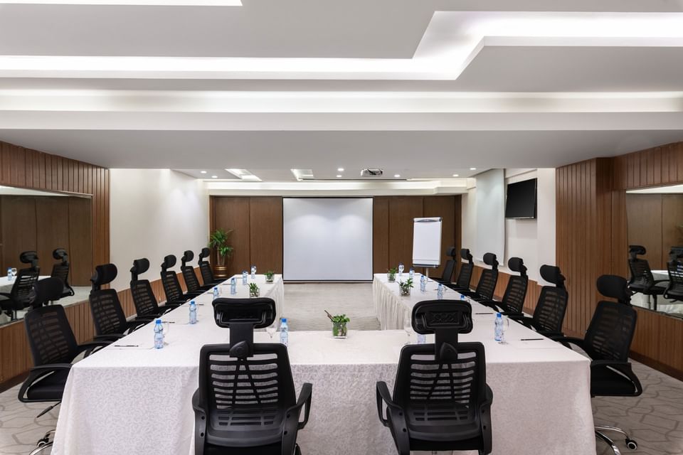 U-shaped table with water bottles by black mesh chairs facing the projector screen in Meeting Room A at Warwick Hotel Jeddah