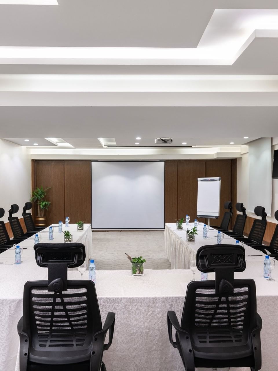 U-shaped table with water bottles by black mesh chairs facing the projector screen in Meeting Room A at Warwick Hotel Jeddah