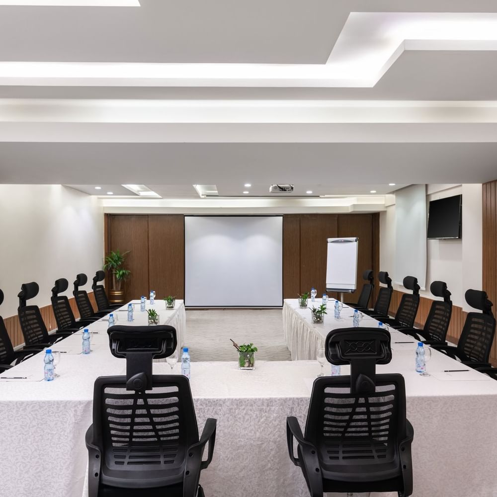 U-shaped table with water bottles by black mesh chairs facing the projector screen in Meeting Room A at Warwick Hotel Jeddah