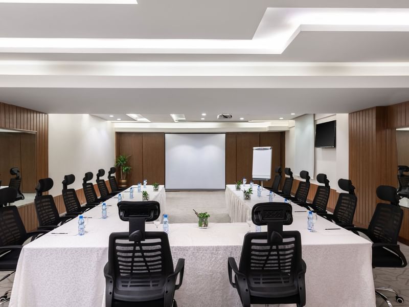 U-shaped table with water bottles by black mesh chairs facing the projector screen in Meeting Room A at Warwick Hotel Jeddah