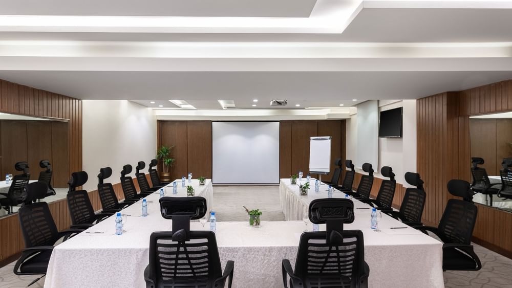 U-shaped table with black mesh chairs facing the projector screen in Meeting Room A at Warwick Hotel Jeddah