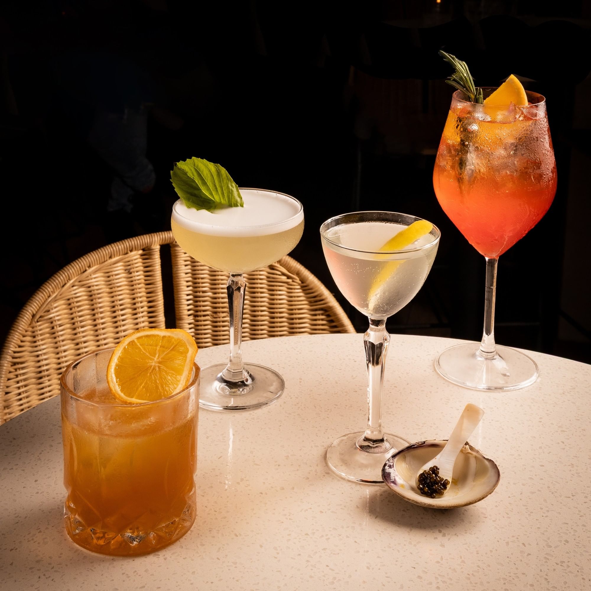 Four artfully garnished cocktails displayed on a white table with a wicker chair and small appetizer plate for Locals Monday.