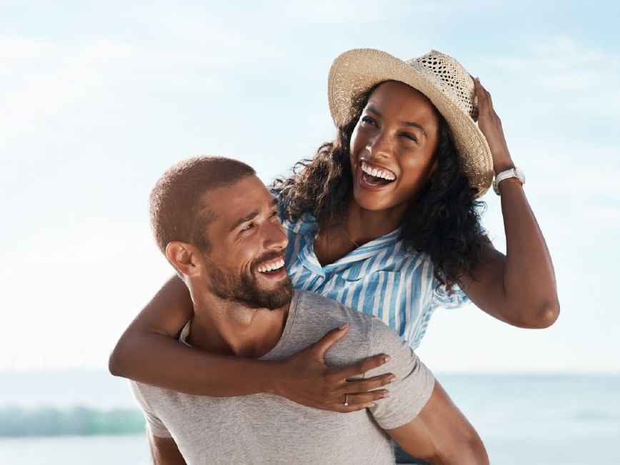 A smiling man lifts a smiling woman in a hat, both with arms around each other, against a beach backdrop.