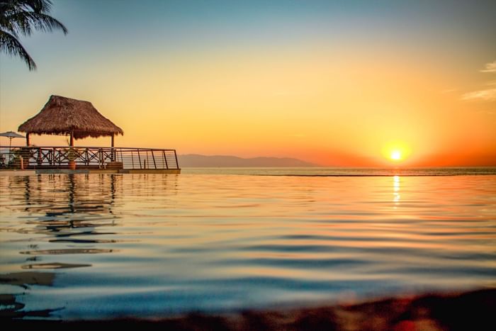Infinity pool reflects a vibrant sunset, with a thatched-roof hut at Puerto Vallarta Resort