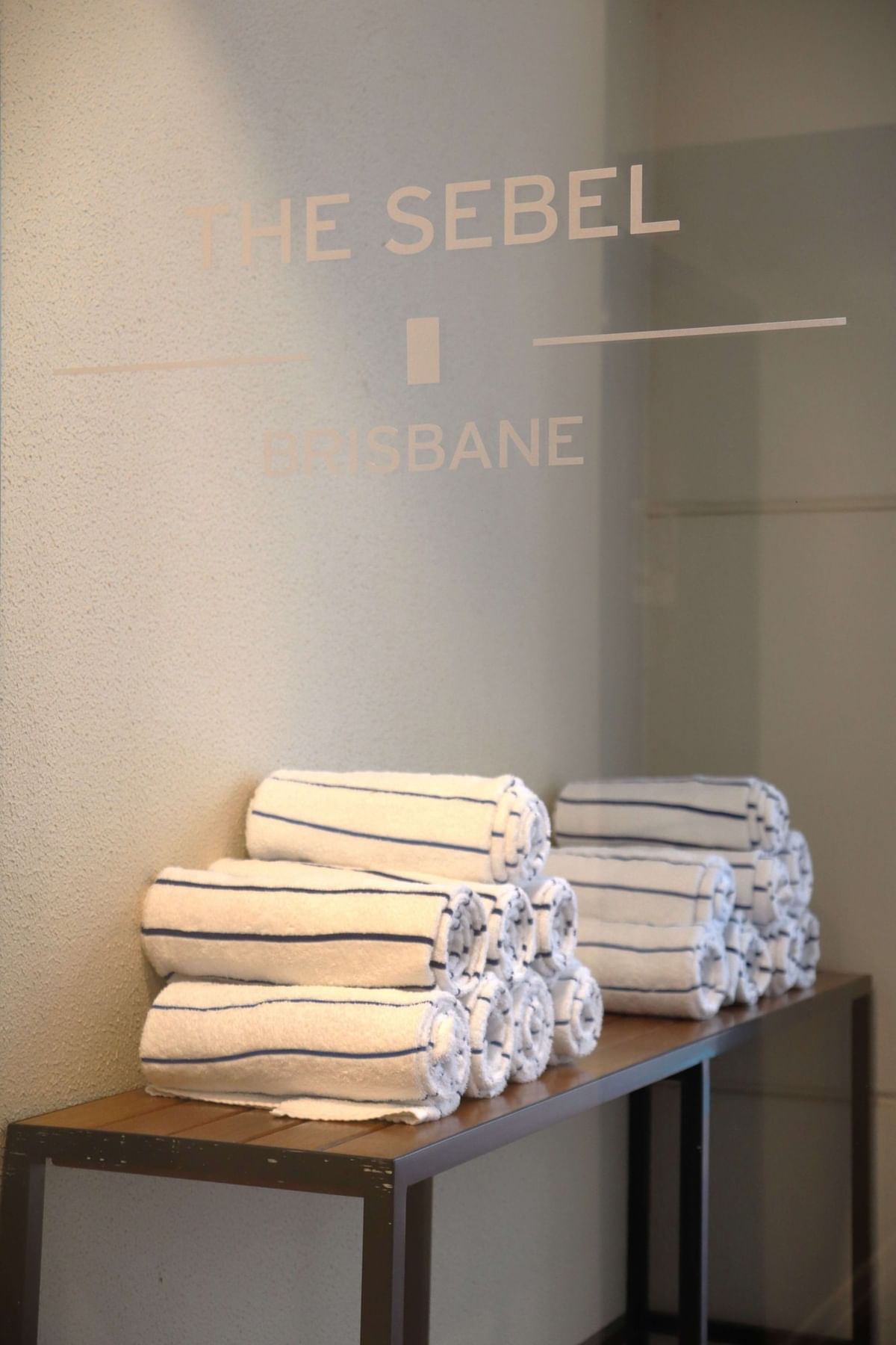 Rolled-up white and blue striped towels rest below a glass panel sign that reads the hotel name at The Sebel Brisbane