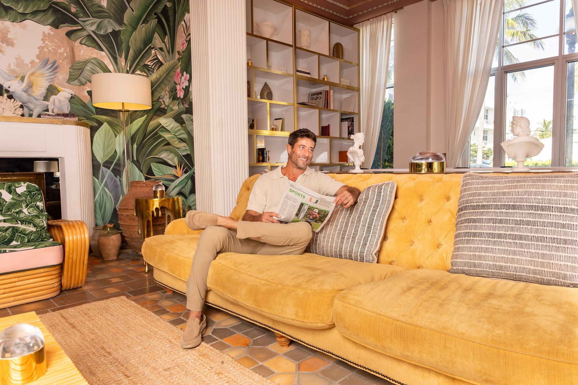 A man sits on a yellow couch in a lobby lounge with a fireplace, bookshelf, and tropical wallpaper.