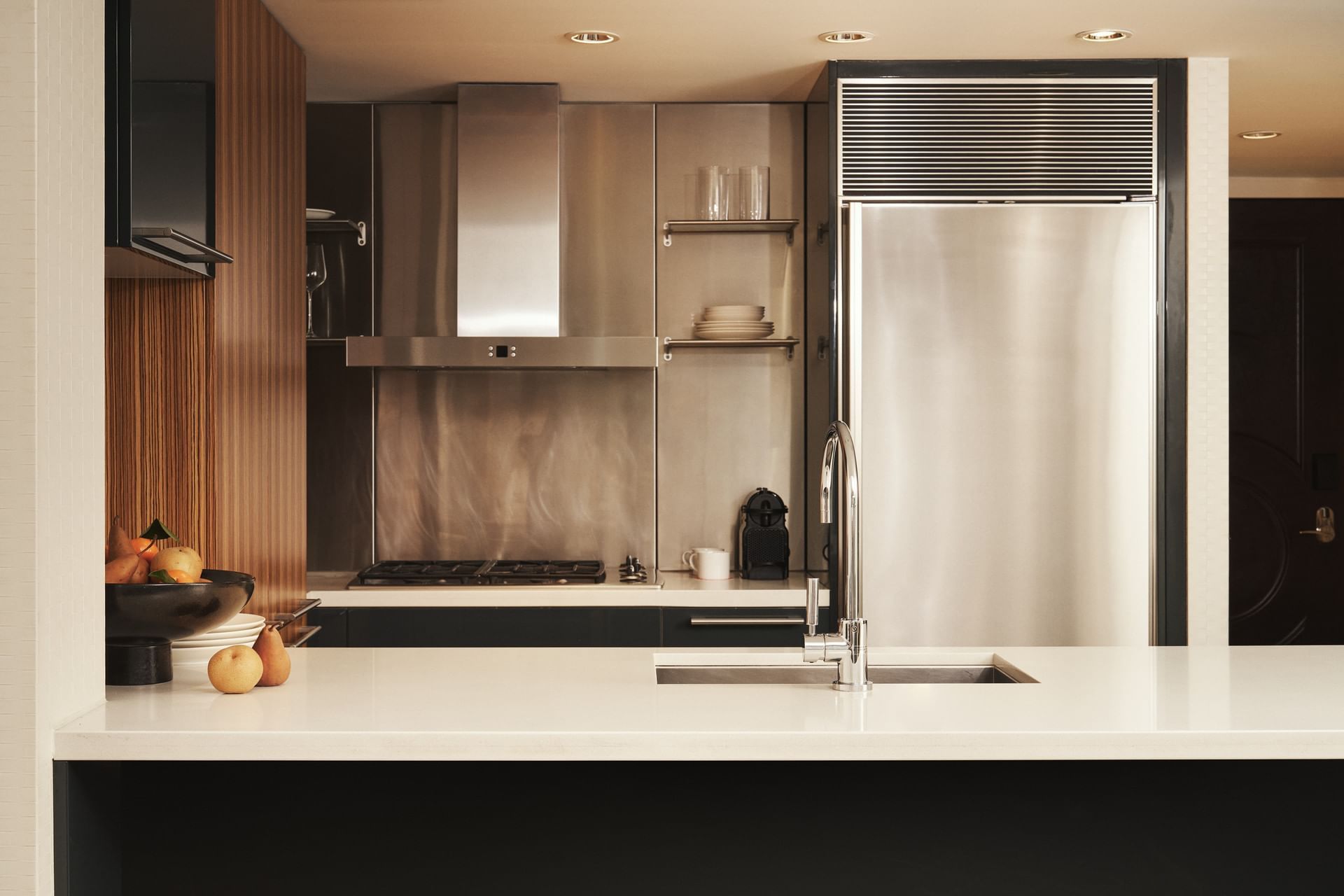 Modern kitchen with white counter, stainless steel appliances, overhead shelf, and fruit bowl on the counter.