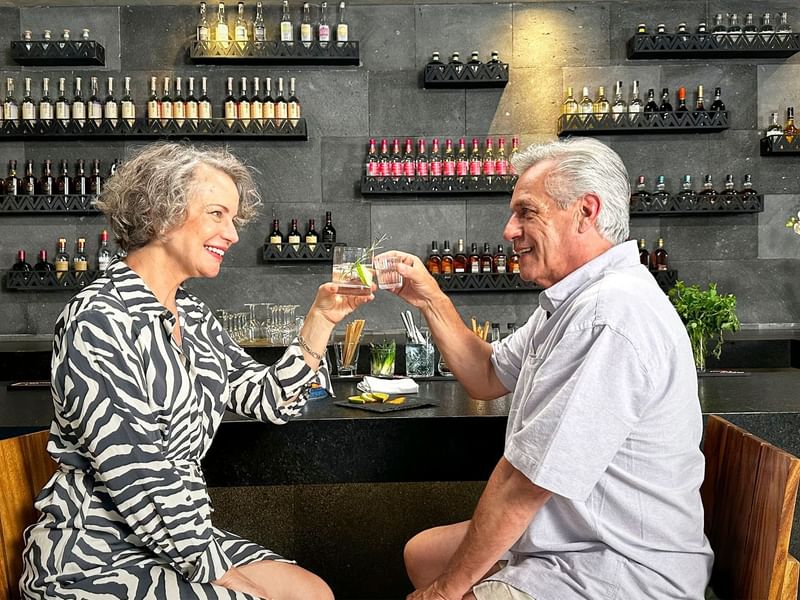 An elderly couple raising glasses in front of a bar at Grand Fiesta Americana