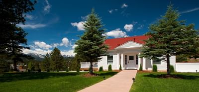 A charming white cottage with a red roof at The Stanley Hotel, surrounded by green grass, pine trees, and a mountain view