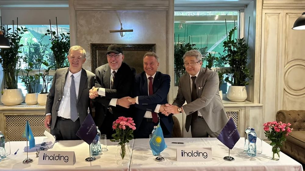 Four business executives shaking hands at a signing ceremony table with flags and flowers in an elegant hotel setting