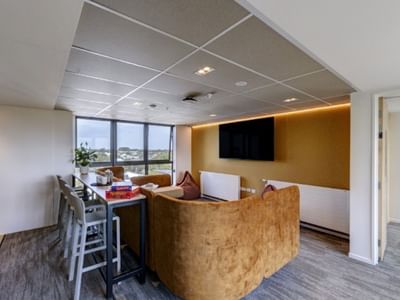 Modern lounge area with tan velvet seating, wall-mounted TV, high-top table, and large windows overlooking the landscape.