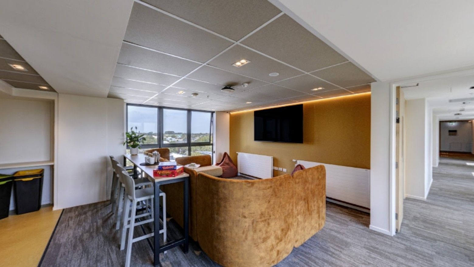 Modern lounge area with tan velvet seating, wall-mounted TV, high-top table, and large windows overlooking the landscape.
