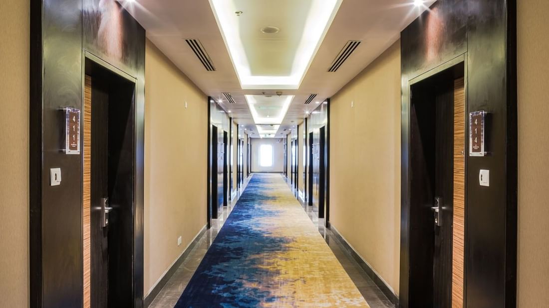 Hallway with a blue and yellow carpet lined with dark wood doors leading to the Standard room at Naviti by Warwick Dammam