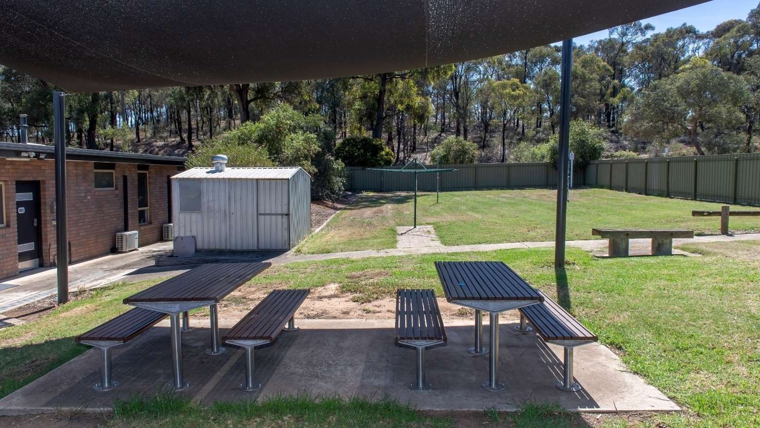 Picnic tables and benches under a shade sail in a grassy courtyard area.
