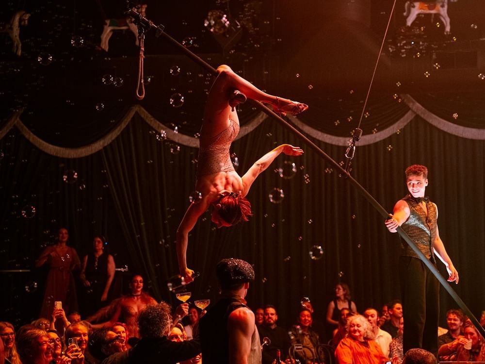 Aerial acrobat performing on trapeze above audience with mime on stage and floating bubbles at Blanc de Blanc Encore.