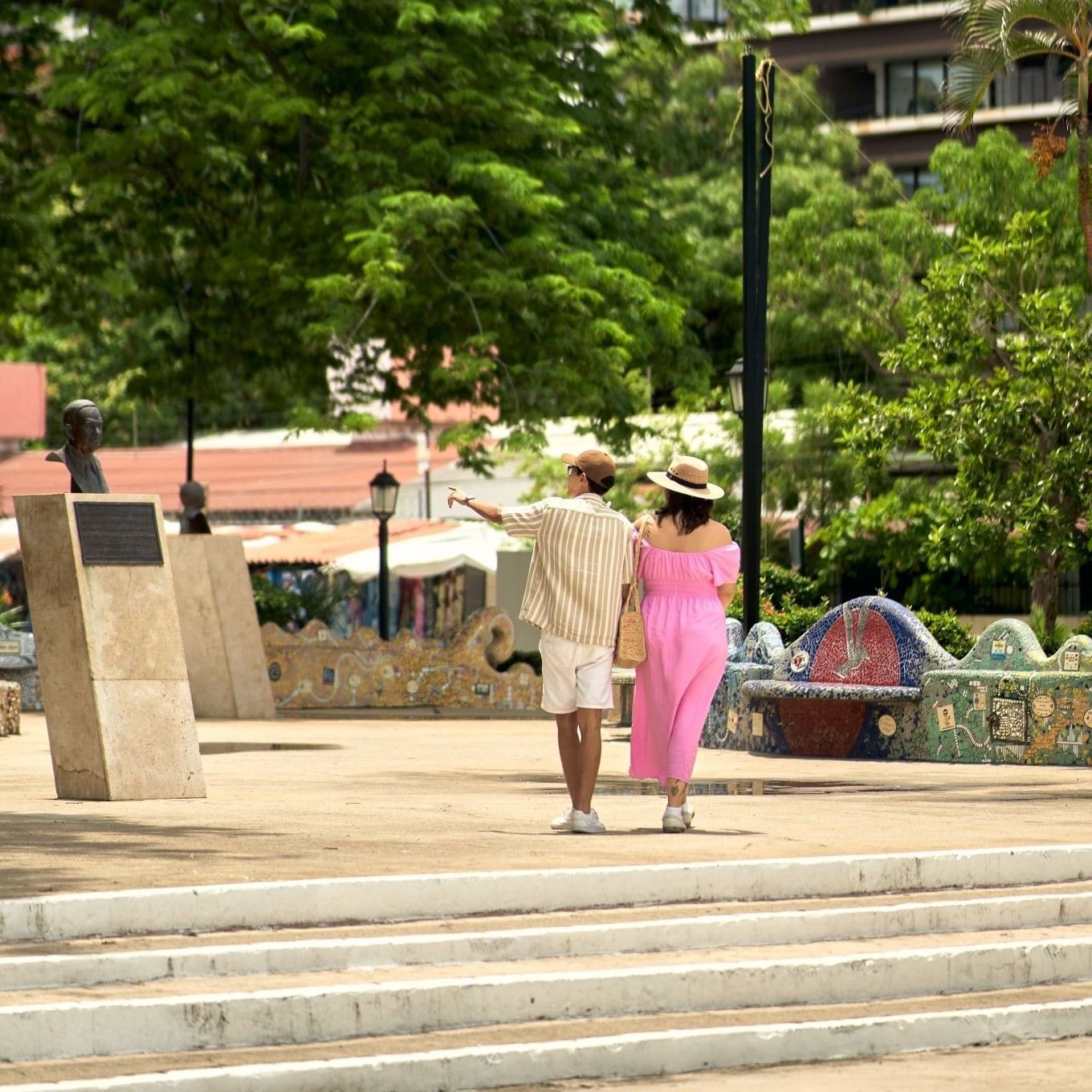 Una pareja caminando por una plaza pública en Puerto Vallarta, con bancas de mosaico coloridas, árboles tropicales y estatuas de bronce bajo la luz brillante del día.