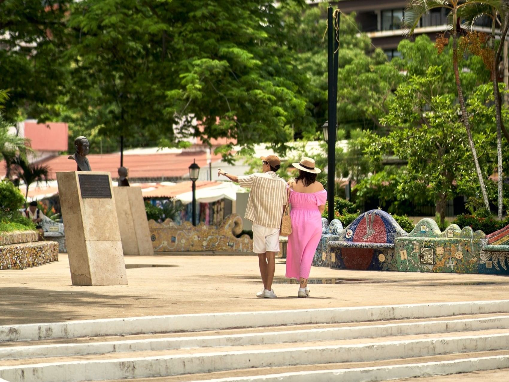 Pareja caminando tomada de la mano en plaza con escaleras, vegetación tropical y edificios al fondo en Puerto Vallarta