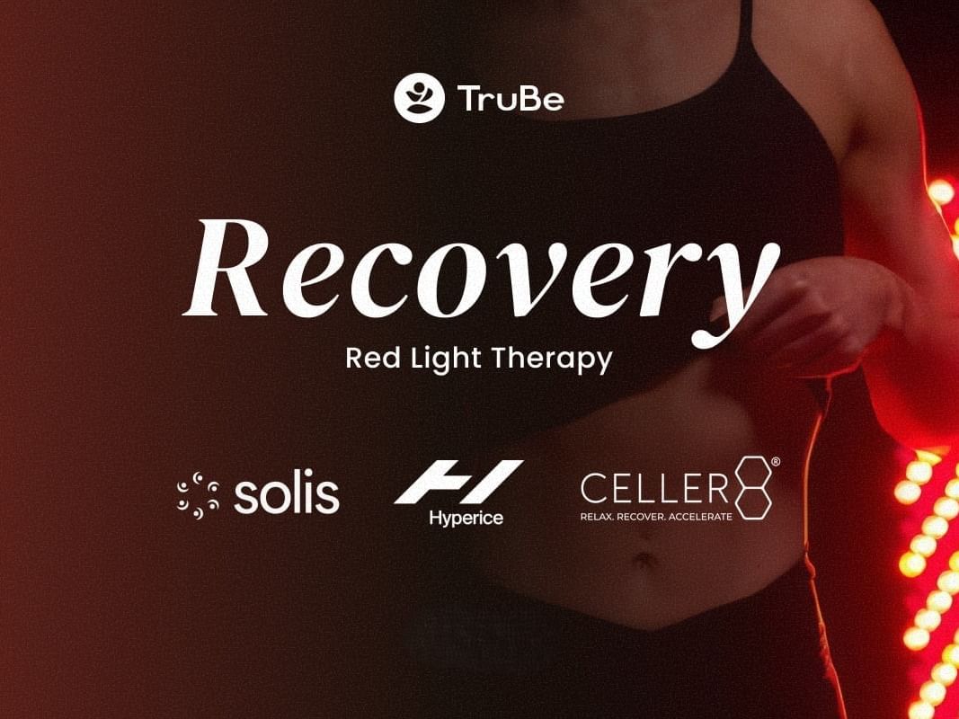 Une personne en tenue de sport se tient debout à côté d'un panneau de luminothérapie rouge dans l'espace bien-être « TruBe Recovery Lounge ».