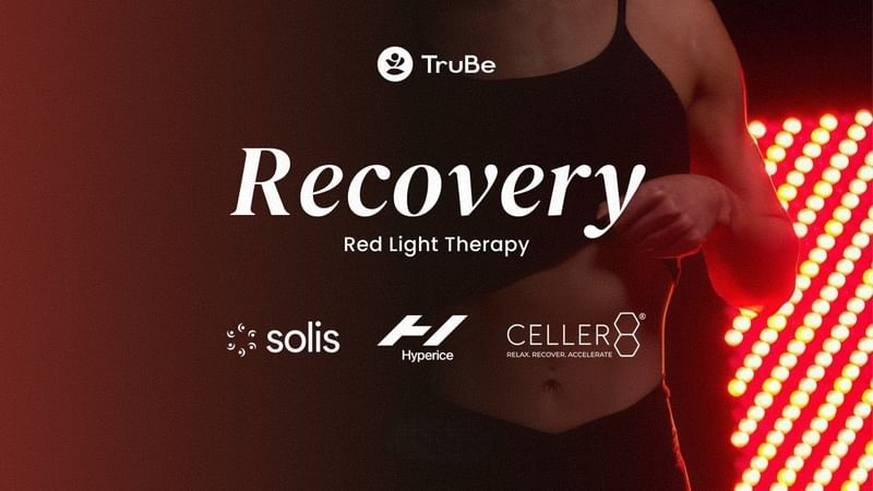 Une personne en tenue de sport se tient debout à côté d'un panneau de luminothérapie rouge dans l'espace bien-être « TruBe Recovery Lounge ».