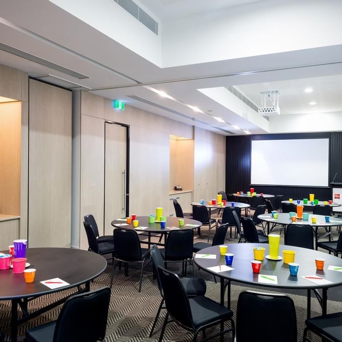 Round tables set up with chairs facing a large projection screen in The Think Tank at Ibis Adelaide