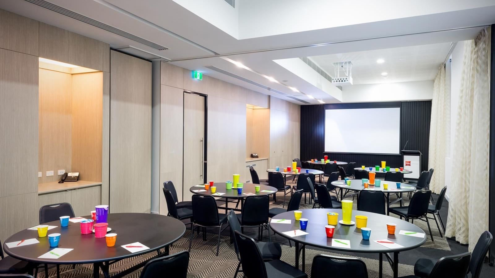 Round tables set up with chairs facing a large projection screen in The Think Tank at Ibis Adelaide