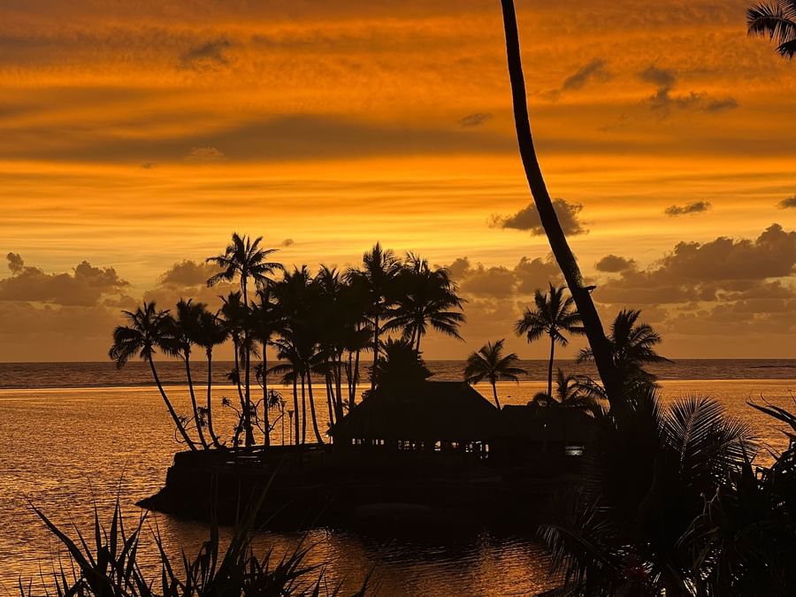 Sunset over ocean with palm trees and huts silhouetted against the sky at Warwick Fiji Resort and Spa.