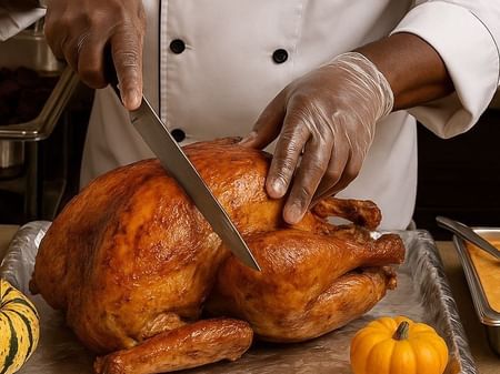 Chef in white uniform carving a roasted turkey on a tray with pumpkins nearby.