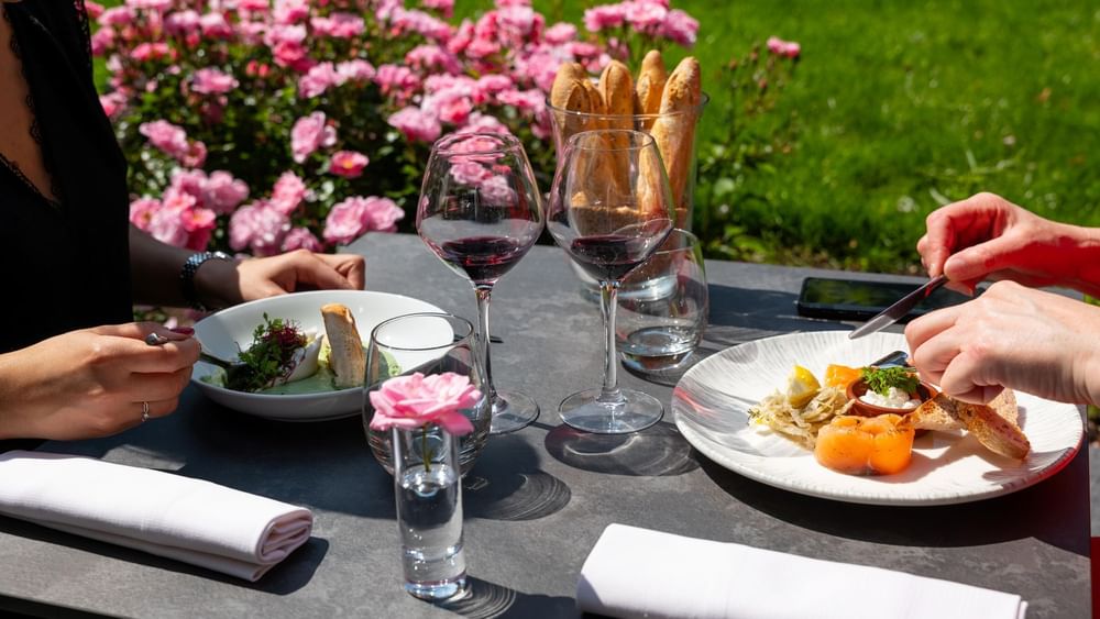 Scène de dîner en plein air mettant en scène deux personnes savourant un repas et du vin rouge chez Warwick Hotels and Resorts, avec des roses roses