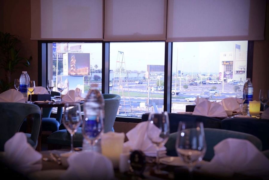 Dining tables with white linens by large windows in Al Multaqa Restaurant at Warwick Al Khobar