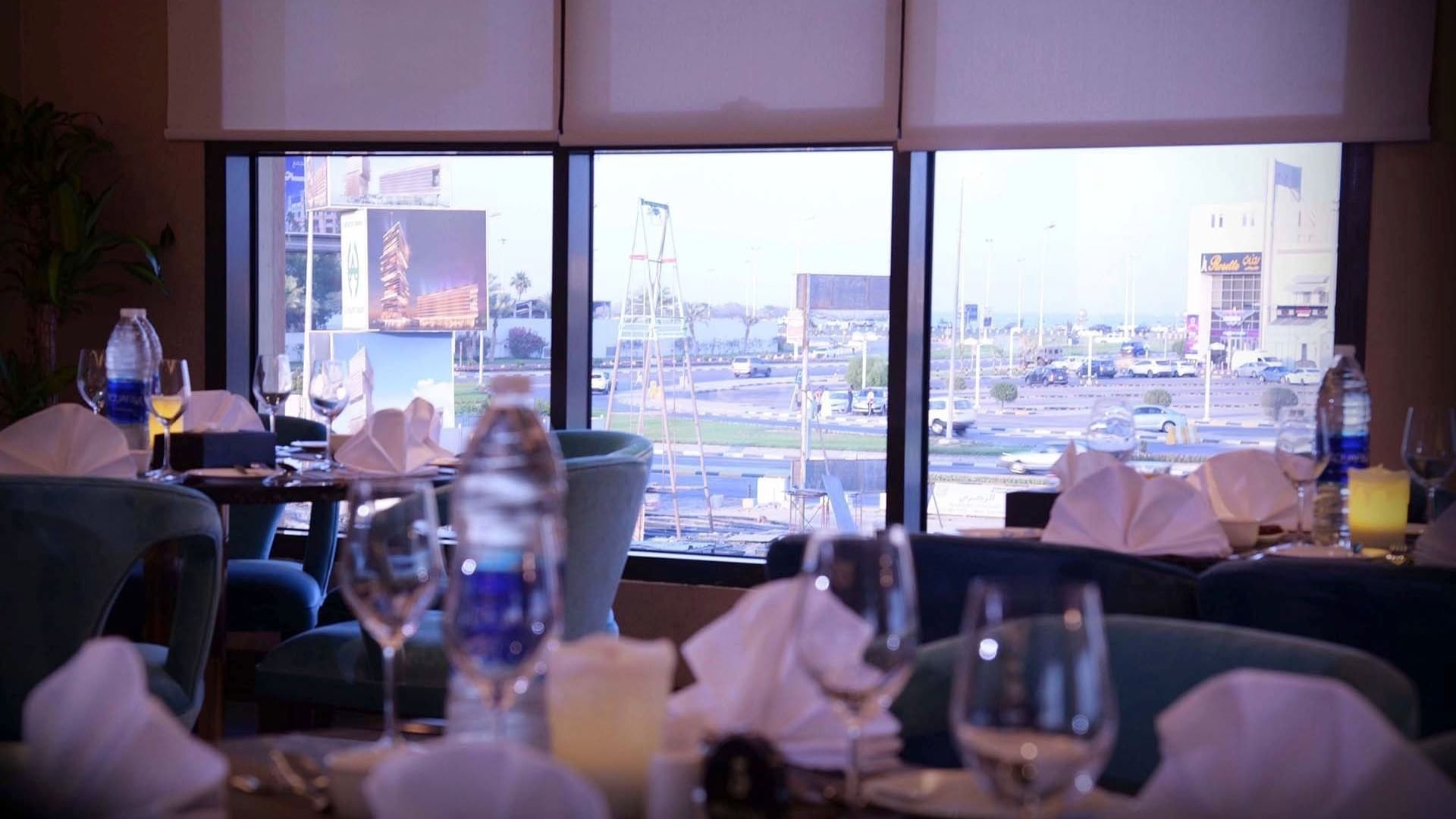Dining tables with white linens by large windows in Al Multaqa Restaurant at Warwick Al Khobar