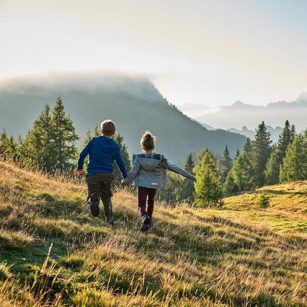 Zwei Kinder laufen Hand in Hand auf einer Wiese mit Bergpanorama im Hintergrund.