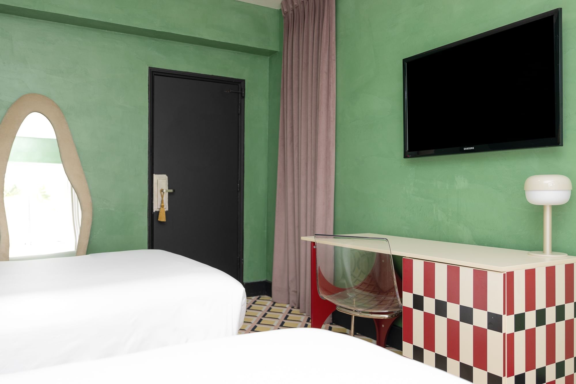A wide view of a hotel room with green walls, a white bed, a checkered desk, and a flat-screen TV