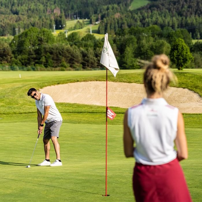Uomo che colpisce una palla da golf, osservato da una donna, vicino a una bandiera.