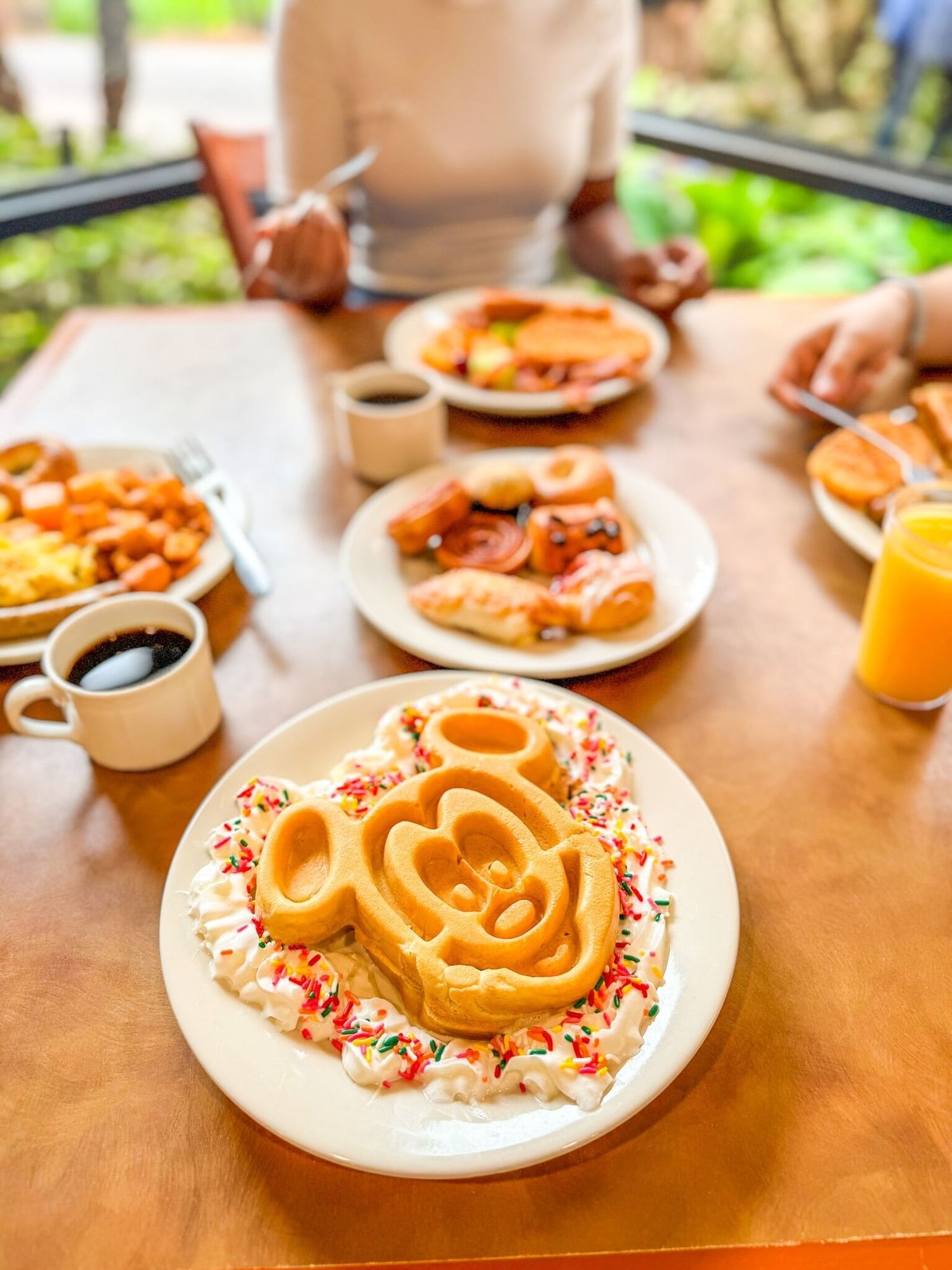 A table with plates of breakfast food, including a large Mickey Mouse-shaped waffle on a bed of whipped cream with sprinkles. 