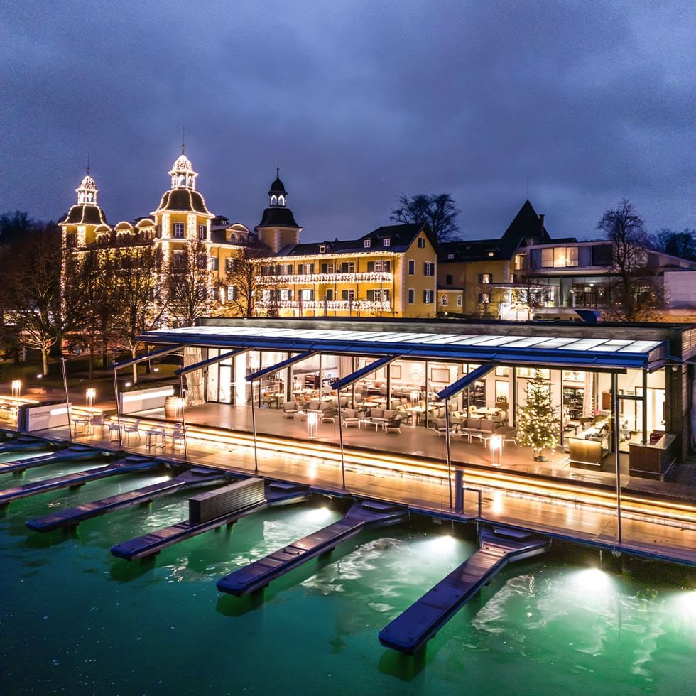 Illuminated waterfront restaurant with dock and large building in background at night.