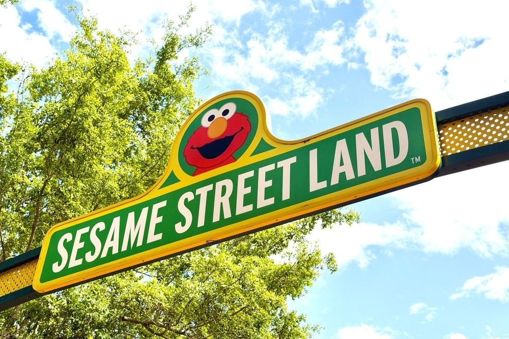 A green and yellow sign reading Sesame Street Land with Elmo's smiling face arches across the sky past a green tree.