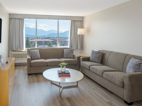 Hotel suite living area with two couches, coffee table, TV, dresser, and large window with mountain view.