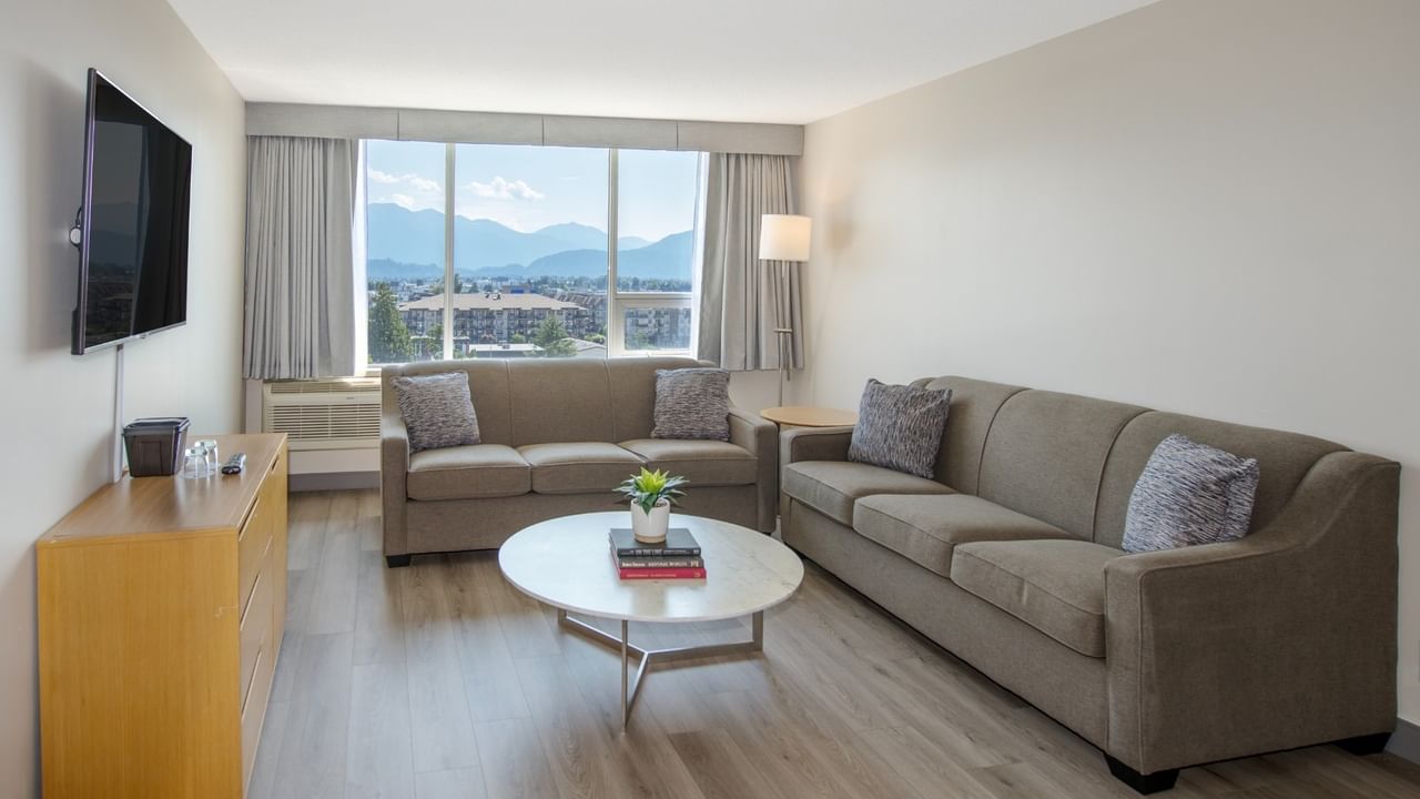 Hotel suite living area with two couches, coffee table, TV, dresser, and large window with mountain view.